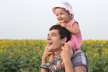 Happy laughing father holding baby daughter on daddy's shoulders standing together in sunflowers meadow enjoying summer walking together on father's day