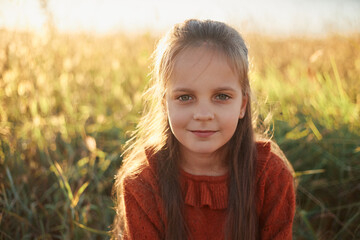 Charming brown haired Caucasian little girl wearing red shirt playing outdoor posing in sunny meadow adorable cute kid looking at camera with gently smile