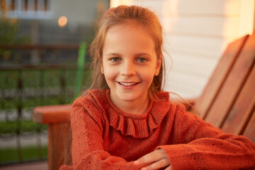 Outdoor portrait of smiling little girl sitting near her house looking at camera with cheerful smile expressing positive emotions relaxing on her terrace
