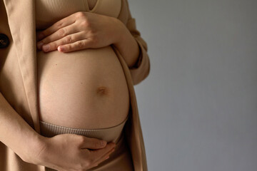 Motherhood, pregnancy, maternity, expectation. Young pregnant woman wearing beige jacket, bra, leggings holding hands on her bare belly, copy space for promotional text