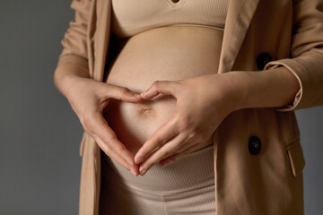 Unrecognizable pregnant woman standing with bare tummy wearing beige jacket making love symbol heart shape with fingers expressing love and tenderness to her expecting baby