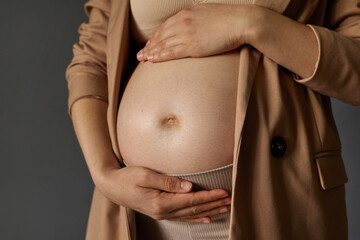 Closeup of young pregnant woman touching her belly and caring about her future child wearing stylish jacket stroking tummy standing isolated over gray background