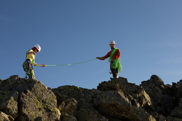 Two people are on a mountain, one of them is holding a rope. The other person is wearing a green...