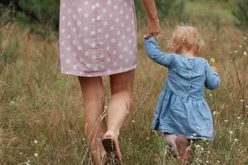Unrecognizable woman legs walking in field on grass holding hand of infant little daughter wearing cute blue dress family walking in meadow on summer day