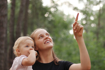 Cute little girl in mother hands looking away while mommy pointing at something interesting in forest young family spending time outdoor on summer day