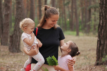 Satisfied cheerful young adult Caucasian woman wearing black casual T-shirt holding baby daughter and embracing older child while enjoying beautiful nature in summer pine trees forest together