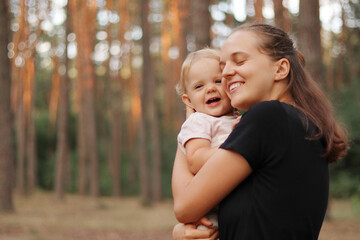 Brown haired cheerful woman hugging with live her cute little daughter in pine tree forest happy mother holding infant kid in her hands enjoying summer walking together