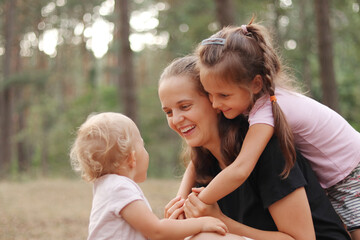 Caucasian woman having fun with live her cute little daughters in pine tree forest happy mother playing with infant kid end elder child enjoying summer weather in wood