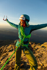 A woman in a green jacket is standing on a mountain with a green rope around her waist. She is smiling and she is enjoying the view - obrazy, fototapety, plakaty
