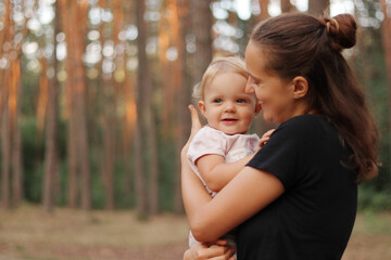 Stylish young mother wearing black T-shirt standing in forest with her infant blond charming...