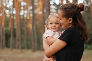 Stylish young mother wearing black T-shirt standing in forest with her infant blond charming daughter family breathing fresh air enjoying summer days on nature