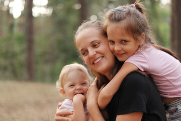 Delighted mother and cute little daughters spending time together on walk in pine tree forest woman...