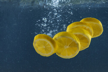 Bright juicy lemon slices with splash and bubbles falling into water isolated over dark blue background, advertisement area