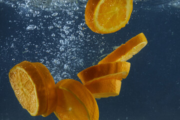 Bright juicy orange slices with splash and bubbles falling into water isolated over dark blue background, advertisement area