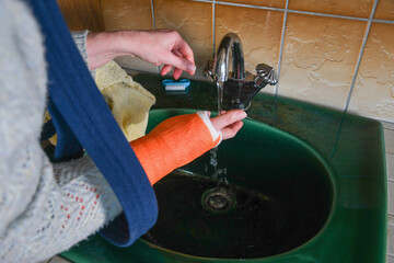  woman with broken arm in cast checks the temperature of the tap water for washing her...