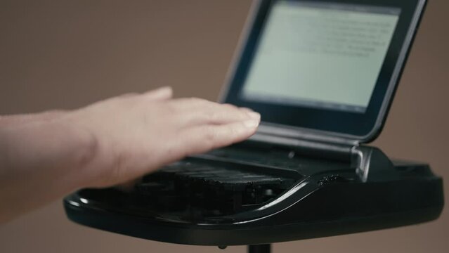 Court reporter typing on stenographer machine with out of focus screen in background