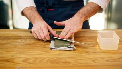 close up a chef in a white jacket in the kitchen at the table sharpens knife on a green stove