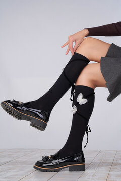 Cropped close-up shot of a woman wearing butterfly decor black knee high socks with a straps. The girl in a brown  long sleeve, a grey skirt, and cotton black knee high socks is posing in a light back