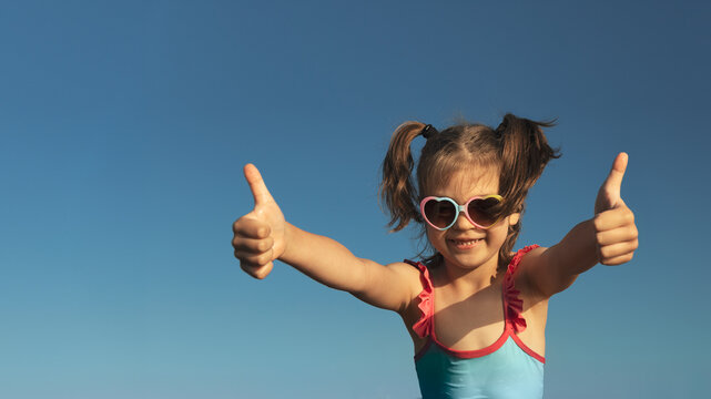 Portrait of laughing girl of 6 years in blue swimsuit and heart-shaped sunglasses gesturing super, fine against blue sky, banner