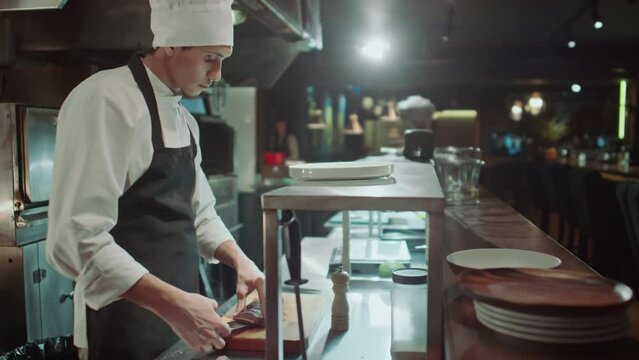 Chef in apron and hat serving cooked food on plate while preparing order in open restaurant kitchenChef in apron and hat serving cooked food on plate while preparing order in open restaurant kitchenCh