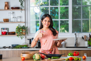 Hppy young asian indian woman in kitchen promoting utensils and ok sign