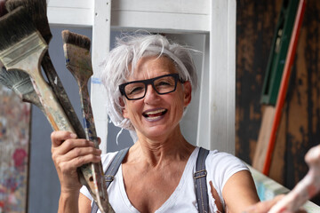 smiling laughing happy older mature woman portrait, proud artist, in her fifties with grey hair and black glasses, with a positive gesture and charisma and many paintbrushes