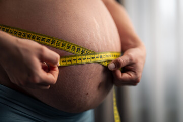 A close-up shot of a pregnant woman using a measuring tape around her round belly - obrazy, fototapety, plakaty
