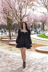 A girl in a stylish black dress walks through a blooming park.