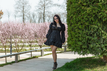 A girl in a stylish black dress walks through a blooming park.