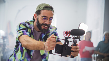 Young Latin Male Filmmaker With Beard And Glasses, Joyfully Operating A Camera In A Busy Studio,...