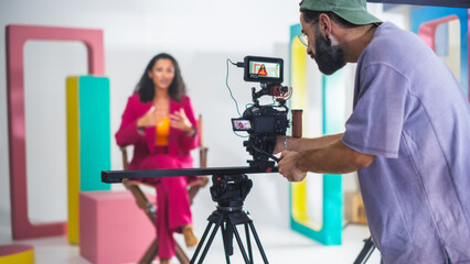 Young Male Cinematographer With Beard, Wearing Cap, Records Vibrant Female Host In Pink Suit During...