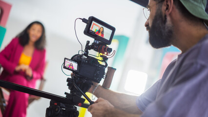 Film Set With A Young Black Female Host In A Pink Suit Engaging In A Lively Discussion, Captured By A Male Cinematographer Focusing Intently On His Camera Equipment, Surrounded By Colorful Backdrops.