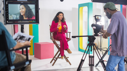 Young Hispanic Woman In A Vibrant Red Suit Speaks Animatedly On A Director's Chair, Surrounded By...