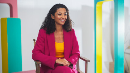Young Black Female Host In Vibrant Pink Suit And Yellow Top, Smiling Engagingly On Colorful Set,...