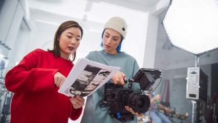 In A Modern Studio, An Asian Woman In A Red Sweater And A Caucasian Woman With Blue Hair, Both Young, Collaborate Over A Camera And Storyboard, Focusing On Creative Film Production And Advertising.