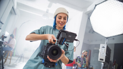 Young Caucasian Female Filmmaker In A Beanie Adjusts A Camera On A Busy Film Set, Capturing The...