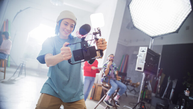 Young Caucasian Female Videographer With Blue Hair, Wearing A Beanie, Captures Content In A Studio, Surrounded By Professional Lighting Equipment And Team Members Engaged In A Creative Production.