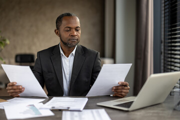 A professional African American businessman attentively reviews paperwork while sitting at his desk in a modern office setup, showing dedication and concentration. - Powered by Adobe