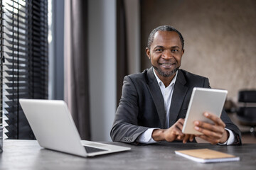 A professional African American businessman engages with a digital tablet at his desk in a modern office setting, portraying confidence and expertise.