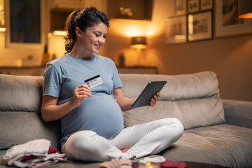 A joyful pregnant woman using a tablet to shop online for baby goods while sitting on the couch in...