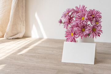 Empty paper card mockup, vase with pink flowers bouquet on beige wooden table, white wall and linen...
