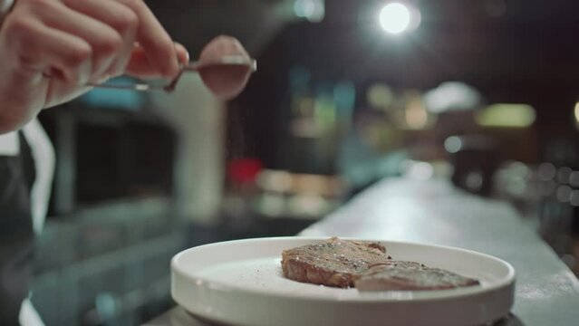 Close up shot of hand of male chef using sifter while adding spices to ready meat steaks served on plate in restaurant kitchenClose up shot of hand of male chef using sifter while adding spices to rea