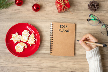 Woman hand writing 2025 PLANS on notebook with Christmas cookies on table. Xmas, Happy New Year, Goals, Resolution, To do list, and Strategy concept