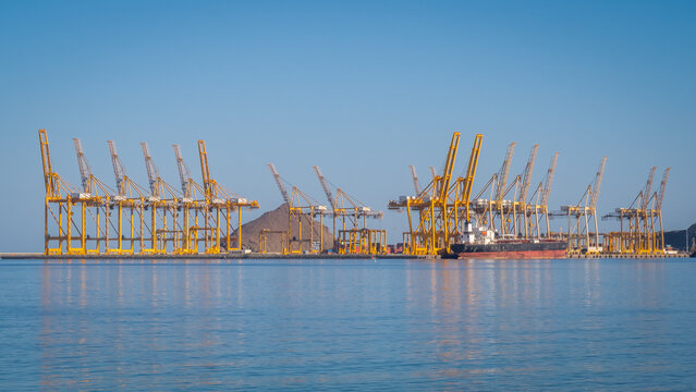 Port cranes loading container ship in  the Port of Fujairah, United Arab Emirates. Rows of yellow port cranes against blue sea and sky.