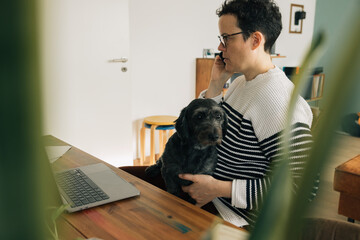 A woman is talking on his cell phone while working at home alongside his little black dog