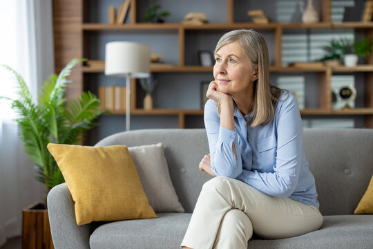A thoughtful senior woman sits alone on a couch, looking away with a reflective expression in a well-furnished living room surrounded by books and plants.
