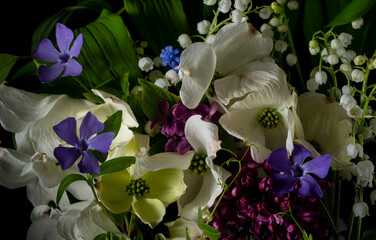 Close up bouquet of beautiful spring flowers on a black background. Dogwood flowers, lilies of the valley, lilac, periwinkle, green leaves. Still life, low key photo.