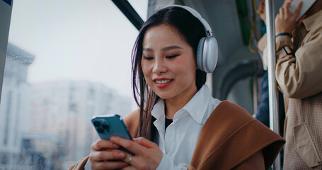Frontal view of Asian girl sitting next to window in train or bus. Woman listening to music in wireless headphones while using smartphone. Talking with friend remotely. Bluetooth connection.