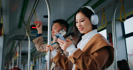 Cute Asian woman standing still next to handsome man. Wearing headphones and stylish coat. Listening to music while scrolling social media on smartphone. Browsing web. Searching for activities.
