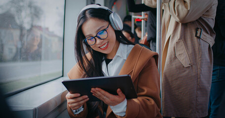 Pretty asian woman sitting next to large window of moving train. Wearing headphones for music while using tablet device. Smiling with joy during trip on public transportation.